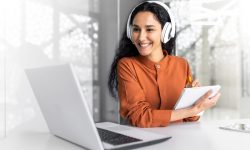 Latin american business woman with curly hair and headphones watching online training course at workplace, woman writing information happy and satisfied with the results of professional development